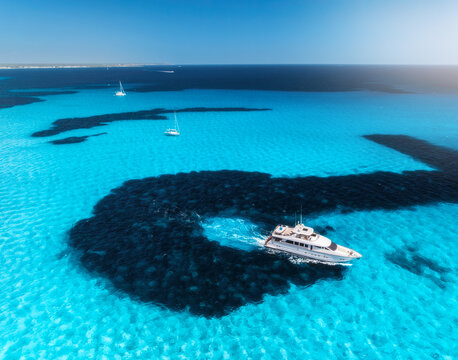 Aerial drone view of luxury yacht moving through shallow turquoise sea above dark rocky seabed, boats anchored nearby, summer scene near Es Trenc, Mallorca, Balearic Islands, Spain. Top view. Seascape