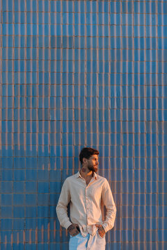 Stylish man with beard and tattoos standing by blue wall