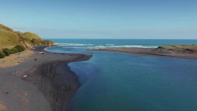 People enjoy a sunny day at Marokopa, Waikato, New Zealand, with some vehicles parked nearby. The scene captures a relaxed atmosphere at a coastal location in New Zealand.