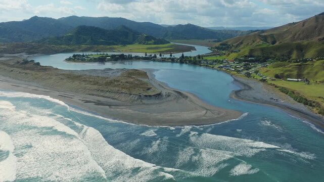 Aerial view of the picturesque coastal village of Marokopa, Waikato, New Zealand, nestled between lush green hills and the Tasman Ocean. The tranquil river mouth meets the sea