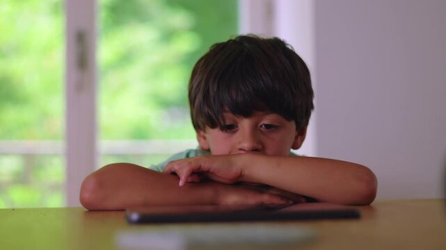 Young boy resting head on folded arms while watching tablet with passive expression showing stillness low energy and absorbed attention during quiet screen time at home