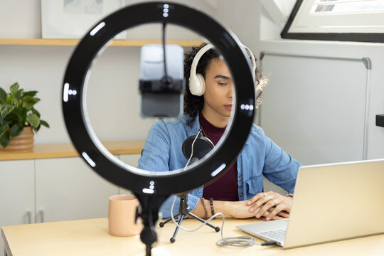 Non-binary person recording podcast at home desk using LED ring light, mic and headphones