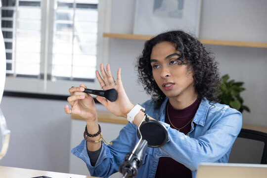Non-binary creator wearing denim jacket showing makeup brush and speaking toward mic at home studio
