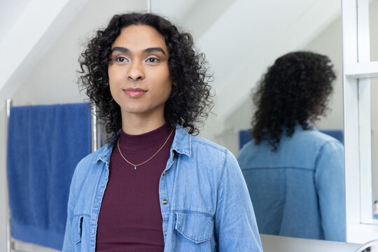 Nonbinary standing in bath wearing denim shirt burgundy top two necklaces mirror blue towel on rack
