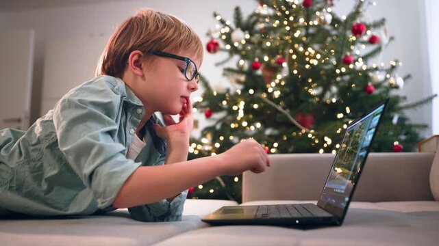 In xmas little boy in glasses lies on sofa with a laptop, thinking then typing