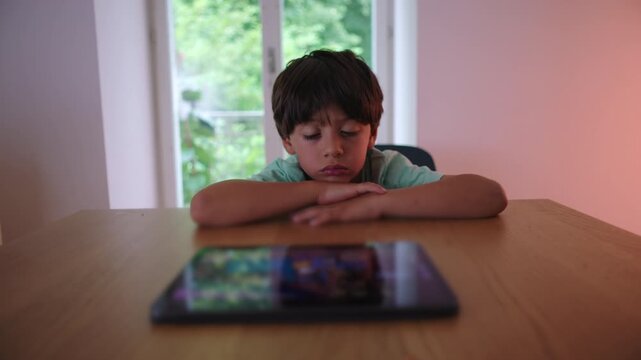 Young boy sitting at table resting chin on hands while watching tablet showing quiet boredom low stimulation and passive engagement with digital content in everyday home setting