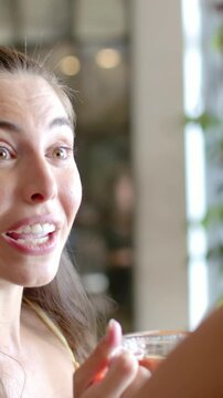 Vertical video: Smiling woman in yellow top reacting to offframe remark at cafe, holding glass cup
