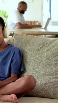 Vertical video: Cam panning son blue T headset tapping pink cell on couch at home dad typing laptop