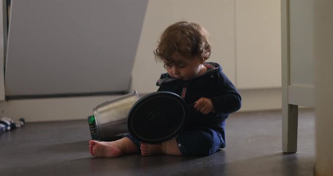 Baby sitting on floor manipulating pot lid and container with both hands, focused posture and downward gaze showing curiosity, coordination and exploratory behavior