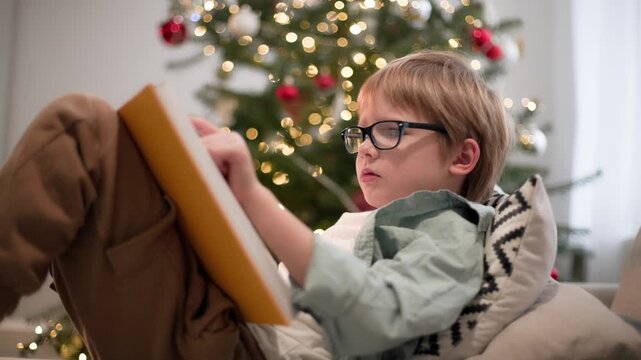 In the bright room, a boy wearing glasses is lying on sofa and reading a book