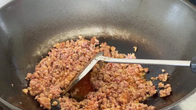 A hand using a spatula to stir fry minced meat in a hot sizzling wok, preparing the base for a dish.