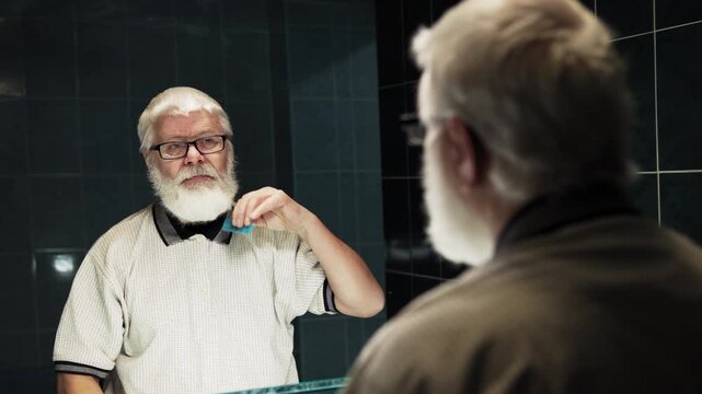 The man in glasses is combing his white hair in bathroom in front of mirror