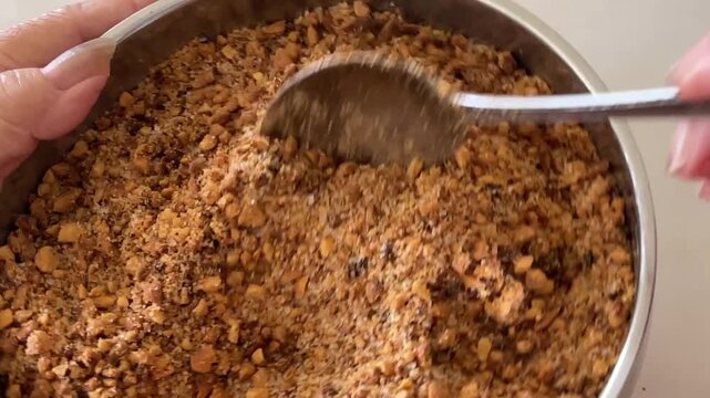 A close-up captures a senior woman's hands using a metal spoon to lightly lift and check a mixture of coarse roasted peanuts and brown sugar, ensuring it is ready for filling.