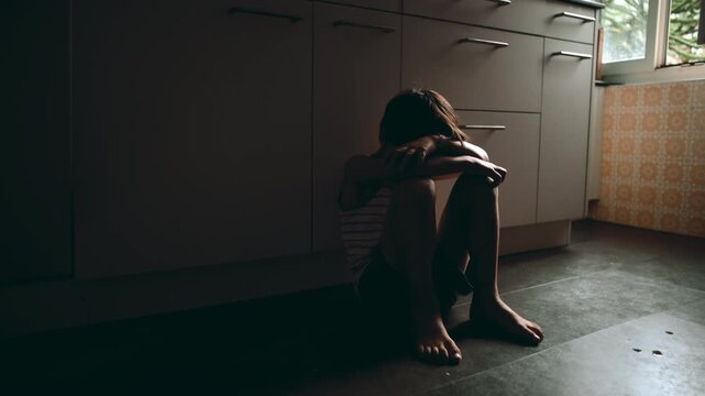 Child sitting alone on kitchen floor in corner hiding face in arms expressing deep loneliness depression and emotional trauma in domestic environment suggesting neglect and inner pain