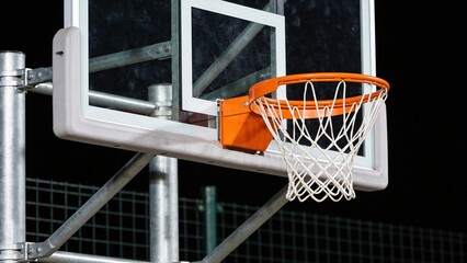 Naklejka premium A basketball hoop with a white net and orange rim is positioned against a dark background. The scene captures the essence of sports and outdoor activities.