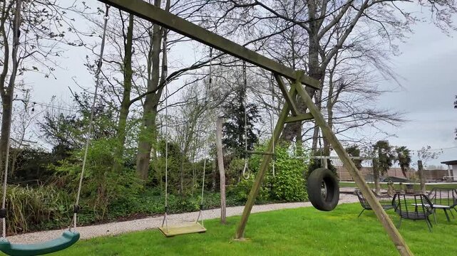 Static shot of an empty wooden swing set on a green lawn.
This horizontal video shows a quiet outdoor playground scene in the Flanders region. A sturdy wooden swing set stands on a well-k