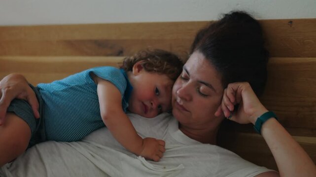 Mother resting on bed with toddler lying across her body showing visible fatigue and emotional weight of caregiving while maintaining gentle connection and protective maternal presence