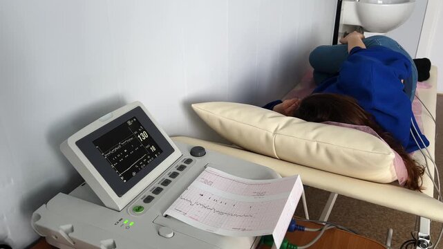 Expectant mother lying on an examination bed during a cardiotocography procedure, with the fetal monitor displaying the baby's heartbeat and printing the results on a graph paper