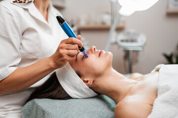 A professional cosmetologist uses a dermapen for a microneedling facial procedure on a young woman lying down in a beauty clinic. © Dusko