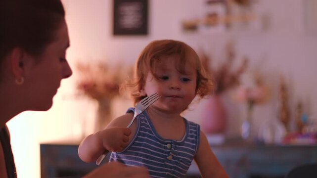 Toddler eating with fork while sitting at table next to mother showing growing independence coordination and self feeding development in everyday family mealtime at home