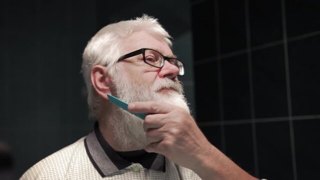 An elderly man combs his white beard, looking at himself in the mirror