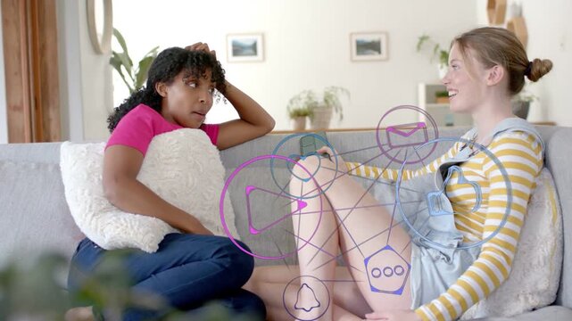 Two teens sitting on gray sofa, right teen tapping phone causing icons aiding marketing