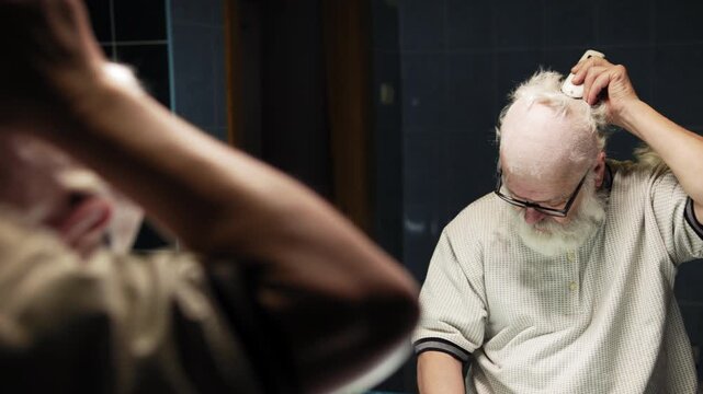 Elderly man in glasses cutting his own hair with a trimmer in front of a mirror