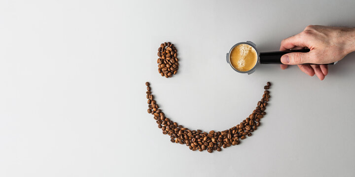 Hand holds portafilter with espresso as eye of smiley made of coffee beans on light background, minimal banner concept with copy space.