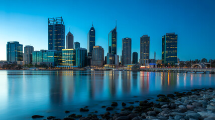Obraz premium A nighttime photograph captures cityscape with buildings, skyscrapers, lights, water, reflection, rocks, and shoreline.