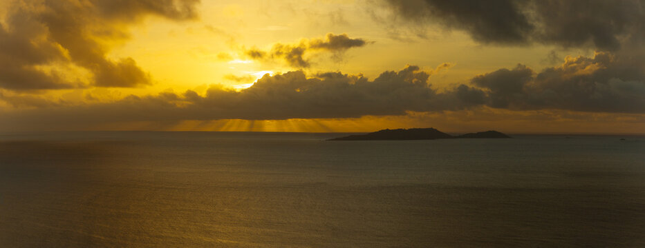Aerial view of the island of Praslin at sunset with golden light rays breaking through dark clouds over the calm Indian Ocean in Seychelles.