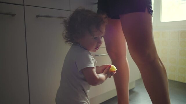 Mother handing apple slice to toddler in kitchen while cooking, gentle caregiving moment, child receiving food, nurturing interaction and everyday parenting routine at home