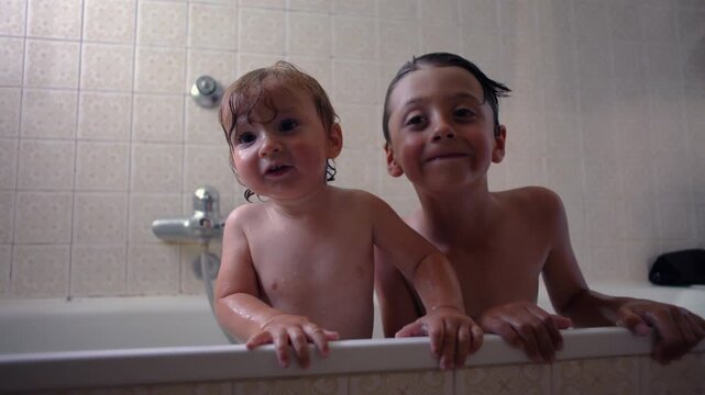 Older child smiling behind baby in bathtub, playful sibling interaction during bath time, warm family moment, hygiene routine, joyful expressions and emotional connection at home