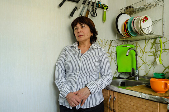 Senior woman standing in kitchen, thoughtful expression near sink, domestic routine concept, mature female at home, casual indoor lifestyle