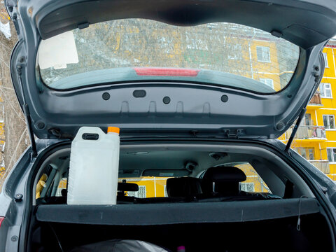 White plastic jerrycan with orange cap on parcel shelf inside open hatchback trunk. Winter city background visible through rear window. Automotive maintenance, emergency supplies, vehicle care