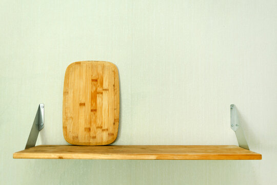 Bamboo cutting board stands on wooden shelf with metal brackets against light wall. Minimalist kitchen storage solution, home organization, cooking preparation, and interior design concept.