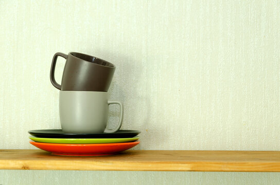 Two stacked mugs and colorful plates arranged on wooden shelf against light wall. Kitchen organization, dishware storage, home decor, tableware arrangement, and interior styling concept.