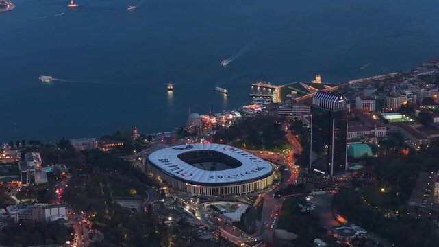 High angle scene of Istanbul at twilight, showing illuminated Vodafone Park stadium, dark waters of Bosphorus Strait with boats, and bustling city streets