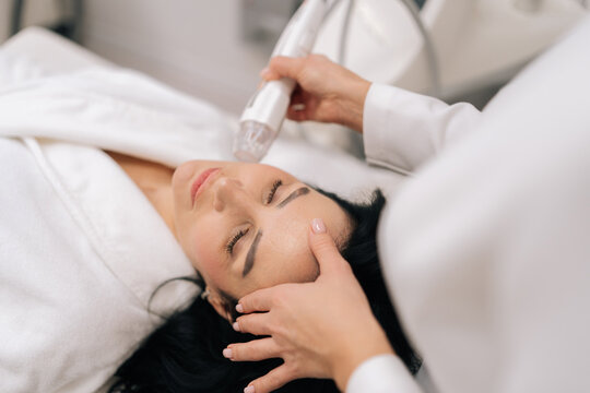 Close-up top view of relaxed woman receiving innovative rf microneedling facelift procedure from skilled esthetician, modern hardware cosmetology method for skin tightening and wrinkle reduction.
