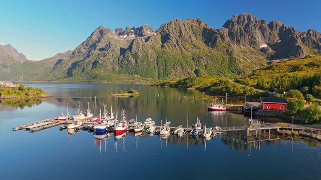  Many boats are docked at the island harbor in the Lofoten Islands, Norway. Drone aerial shot shows sharp mountains reflecting in the calm fjord water of the Lofoten Islands, Norway