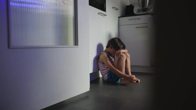 Child isolated in kitchen corner with body curled inward and face hidden expressing severe emotional distress depression and childhood trauma within silent domestic environment