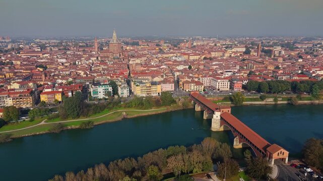 The historic Ponte Coperto spans across the Ticino River in Pavia, Italy. The Duomo di Pavia rises above the residential streets and red roofs of Pavia, Italy. 4K drone aerial
