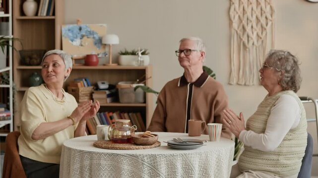 Medium long stab shot of female Caucasian caregiver bringing out cake with candles to elderly man celebrating birthday with female friends sitting at table in nursing home