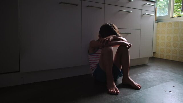 Child sitting on kitchen floor near window with head lowered and arms covering face expressing emotional exhaustion loneliness and mental health struggle in domestic space