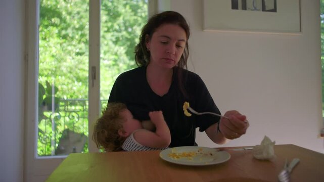 Mother breastfeeding toddler at table while eating meal showing multitasking caregiving and natural everyday parenting moment in home environment