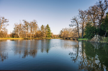 Obraz premium Scenic view of a calm lake with reflections of trees and a wooden bridge in Lednice Castle park. Beautiful sunny spring day in the South Moravia region, Czech Republic.