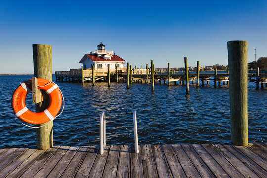 A sunset landscape of the wooden dock with an orange life preserver and the Roanoke Marshes Lighthouse in the background. Manteo, North Carolina