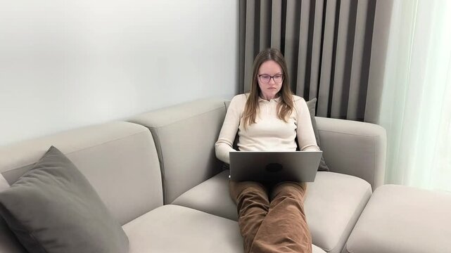 Woman works on laptop sitting on sofa at home interior. Female uses computer typing during remote activity with focused attention. Productivity concept showing freelance digital occupation