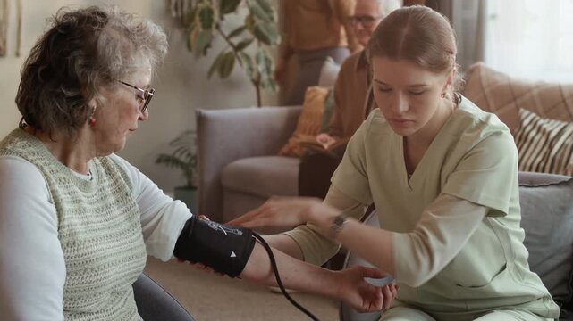 Medium long handheld shot of female Caucasian caregiver in uniform measuring blood pressure of senior woman while working in nursing home