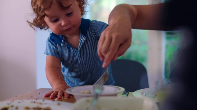 Toddler eating with fork independently while mother supports nearby, natural mealtime moment showing growing autonomy, coordination, and guided learning in everyday family routine