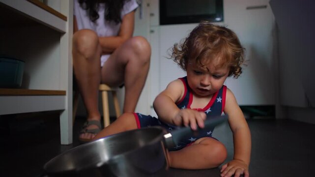 Toddler sitting on kitchen floor holding metal pot and tilting it, focused expression, parent nearby, playful exploration of household objects, early learning and curiosity at home